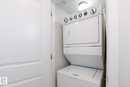 White stacked laundry appliances within a dedicated utility closet, illuminated by a flush-mount ceiling light - #110 9523 160 Av Nw, Edmonton, AB - Indoor Photo Showing Laundry Room