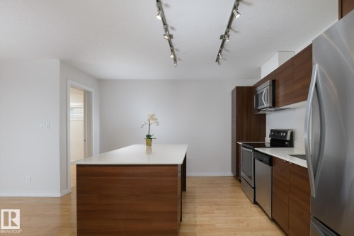 Kitchen featuring wood-finish cabinetry, stainless steel appliances, a white solid surface island, track lighting, and wood-finish flooring - #110 9523 160 Av Nw, Edmonton, AB - Indoor Photo Showing Kitchen With Stainless Steel Kitchen