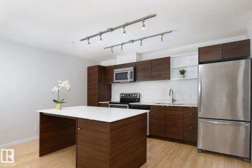 Contemporary kitchen featuring wood-finish cabinetry, stainless steel appliances, white countertops, and a central island with integrated seating - #110 9523 160 Av Nw, Edmonton, AB - Indoor Photo Showing Kitchen With Stainless Steel Kitchen