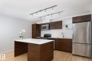 Contemporary kitchen featuring wood-finish cabinetry, stainless steel appliances, white countertops, and a central island with integrated seating - #110 9523 160 Av Nw, Edmonton, AB 