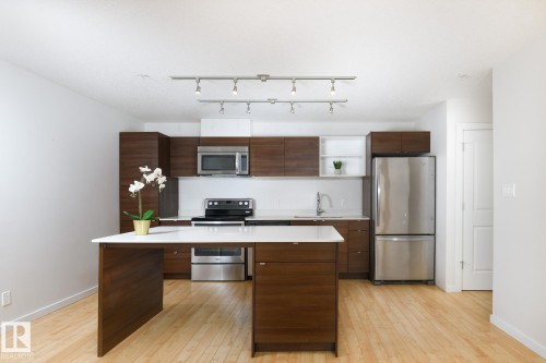 Kitchen featuring wood-finish cabinetry, stainless steel appliances, a center island with a white countertop, and track lighting - #110 9523 160 Av Nw, Edmonton, AB - Indoor Photo Showing Kitchen With Stainless Steel Kitchen