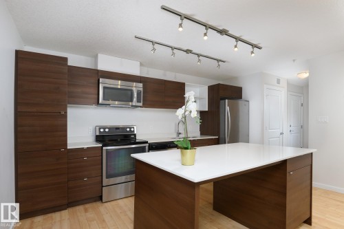 Kitchen featuring wood-finish cabinetry, stainless steel appliances, track lighting, light wood-finish flooring, and a central island with a solid surface countertop - #110 9523 160 Av Nw, Edmonton, AB - Indoor Photo Showing Kitchen