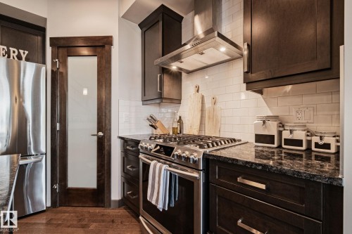 Stainless steel gas range with coordinating hood, dark wood-finish cabinetry, granite-style countertops, subway tile backsplash, and wood-finish flooring - 1337 Secord Landing Nw, Edmonton, AB - Indoor Photo Showing Kitchen With Stainless Steel Kitchen With Upgraded Kitchen
