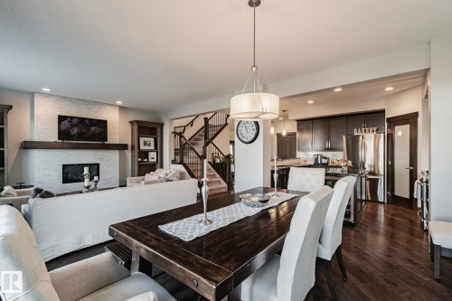 Open-concept design featuring wood-finish flooring, a contemporary fireplace with a stone surround and wood mantle, a staircase with dark wood railings, and a kitchen with dark cabinetry and stainless steel appliances - 1337 Secord Landing Nw, Edmonton, AB - Indoor Photo Showing Other Room With Fireplace