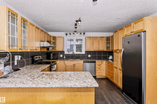Spacious kitchen featuring wood-finish cabinetry, granite-style countertops, dark tile backsplash, stainless steel appliances, and dark wood-look flooring - 6016 177 Street Nw, Edmonton, AB - Indoor Photo Showing Kitchen With Stainless Steel Kitchen