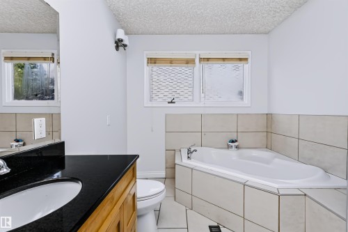 Corner soaking tub with tiled surround, wood-finish vanity with an undermount sink, and black countertop - 6016 177 Street Nw, Edmonton, AB - Indoor Photo Showing Bathroom