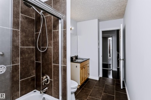 Bathroom featuring a shower-tub combination with dark tile surround, glass shower enclosure, single vanity with wood-finish cabinetry, dark countertop, and dark tile flooring - 6016 177 Street Nw, Edmonton, AB - Indoor Photo Showing Bathroom