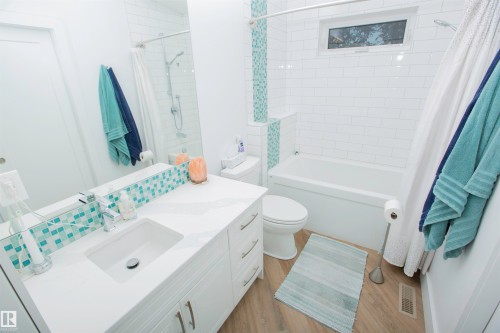 Bathroom featuring a white subway tile shower surround with an integrated accent tile stripe, a white vanity with an integrated sink, a large vanity mirror, and wood-finish flooring - 11131 52 Street Nw, Edmonton, AB - Indoor Photo Showing Bathroom