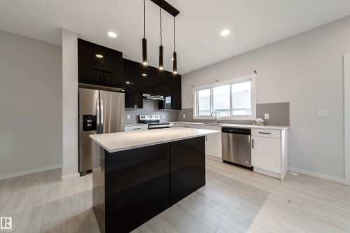 Modern kitchen featuring a central island with a light-toned countertop, stainless steel appliances, and dark-toned high-gloss cabinetry - 7109 Chivers Close Sw, Edmonton, AB - Indoor Photo Showing Kitchen With Stainless Steel Kitchen