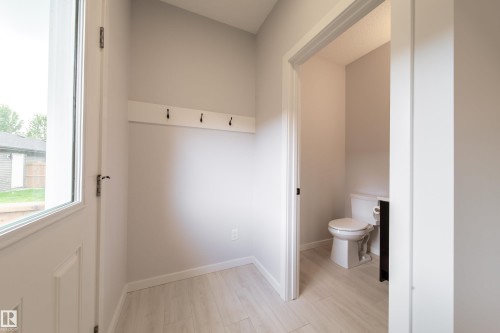 Light-toned wood-finish flooring, white wall paneling with black coat hooks, and a white interior door with a partial glass insert - 7109 Chivers Close Sw, Edmonton, AB - Indoor Photo Showing Bathroom