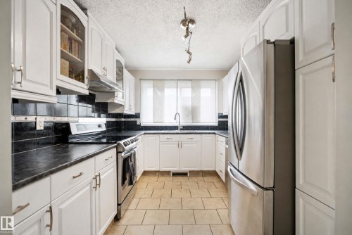 White cabinetry with brushed nickel hardware, dark countertops, a black tile backsplash, stainless steel appliances, and tiled flooring - #25 3811 85 St Nw, Edmonton, AB - Indoor Photo Showing Kitchen