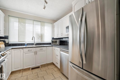 Kitchen featuring white cabinetry, stainless steel appliances, dark countertops, a dark tile backsplash, and light-toned floor tiles - #25 3811 85 St Nw, Edmonton, AB - Indoor Photo Showing Kitchen
