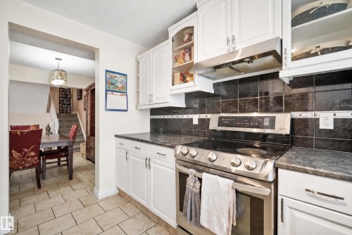 White cabinetry, black tile backsplash, stainless steel range with overhead vent, and dark countertops - #25 3811 85 St Nw, Edmonton, AB - Indoor Photo Showing Kitchen