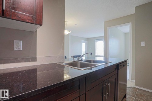Kitchen featuring dark wood-finish cabinetry, a dual-basin stainless steel sink, dark polished countertops, and a built-in dishwasher - #321 270 Mcconachie Dr Nw, Edmonton, AB - Indoor Photo Showing Kitchen With Double Sink