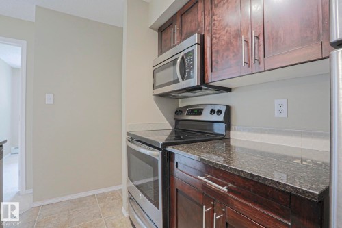 Dark wood-finish cabinetry with silver-tone hardware, stainless steel appliances, speckled countertop, and tiled flooring - #321 270 Mcconachie Dr Nw, Edmonton, AB - Indoor Photo Showing Kitchen