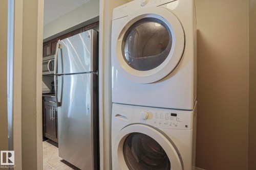 Integrated stackable washer and dryer, stainless steel refrigerator, built-in microwave, and wood-finish cabinetry - #321 270 Mcconachie Dr Nw, Edmonton, AB - Indoor Photo Showing Laundry Room