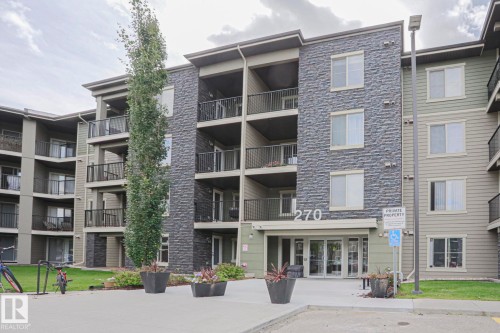 Modern multi-story building featuring stone veneer accents, individual balconies with black railings, a paved entrance, and a landscaped perimeter with mature trees and planters - #321 270 Mcconachie Dr Nw, Edmonton, AB - Outdoor With Balcony With Facade