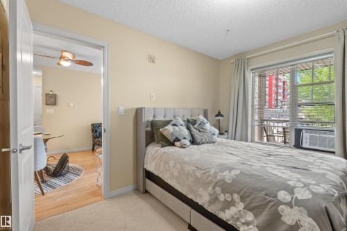 Carpeted flooring, window with blinds, window-mounted air conditioning unit, and a doorway opening to wood-finish flooring - #209 103 Ambleside Drive Sw, Edmonton, AB - Indoor Photo Showing Bedroom