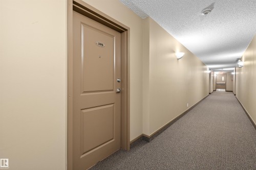 Building hallway with textured carpet flooring, light neutral-toned walls, and sconce lighting fixtures - #209 103 Ambleside Drive Sw, Edmonton, AB - Indoor Photo Showing Other Room