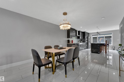 Open-concept dining area with a contemporary chandelier, tile flooring, and a sightline to the kitchen featuring glossy dark cabinetry, an island with seating, and stainless steel appliances - 9329 Connors Road Nw, Edmonton, AB - Indoor Photo Showing Dining Room