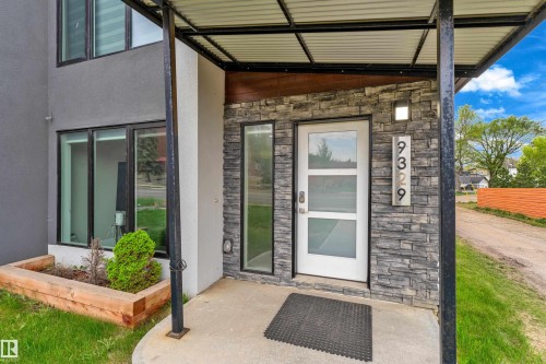 Modern exterior entrance featuring stacked stone accent wall, white entry door with frosted glass panels, corrugated metal porch awning, contemporary address numbering, and integrated landscaping planter - 9329 Connors Road Nw, Edmonton, AB - Outdoor With Exterior