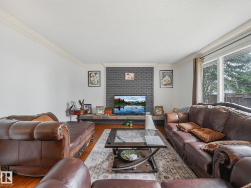 Elegant living room showcasing rich wood flooring, a brick fireplace accent wall with integrated television, and abundant natural light from expansive windows - 10912 5 Avenue Sw, Edmonton, AB - Indoor Photo Showing Living Room