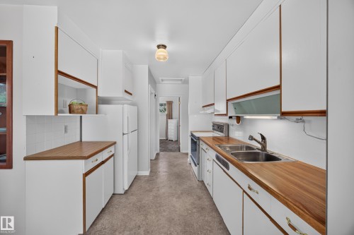 Galley kitchen with white cabinetry, wood-finish countertops, tile backsplash, and dual basin stainless steel sink - 5207 48 Avenue, Leduc, AB - Indoor Photo Showing Kitchen With Double Sink