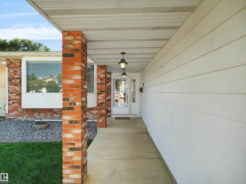 Welcoming covered entry featuring distinctive brick pillars and columns, white horizontal siding, and a manicured lawn with mature landscaping - 7357 18 Avenue Nw, Edmonton, AB - Outdoor With Exterior