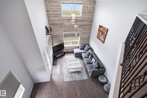 Vaulted ceiling, wood-finish flooring, two large windows, linear pendant lighting, and an open wrought iron railing - #2415 9357 Simpson Dr Nw, Edmonton, AB - Indoor Photo Showing Other Room