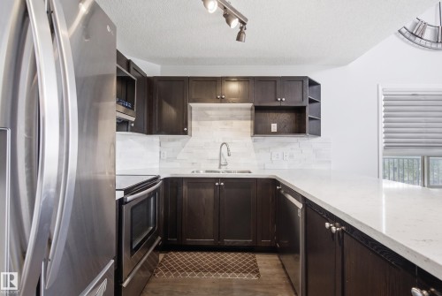 Kitchen featuring stainless steel appliances, dark wood-finish cabinetry, light-toned countertops, and a white tile backsplash - #2415 9357 Simpson Dr Nw, Edmonton, AB - Indoor Photo Showing Kitchen With Stainless Steel Kitchen With Upgraded Kitchen