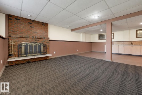 Spacious basement recreation room featuring exposed brick fireplace with decorative surround, neutral taupe wall paneling, patterned carpet flooring, recessed ceiling lighting, and abundant square footage ideal for entertaining or flexible living space - 6768 39 Avenue Nw, Edmonton, AB - Indoor With Fireplace