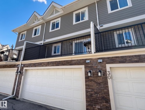 Multi-level exterior featuring horizontal siding, stacked stone facade, white garage doors, and a balcony with black railings - #29 1150 Windermere Wy Sw, Edmonton, AB 