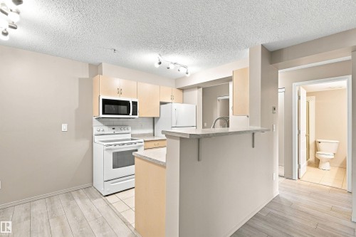 Kitchen with light wood-finish cabinetry, white appliances, and a breakfast bar - #117 17011 67 Av Nw, Edmonton, AB - Indoor Photo Showing Kitchen