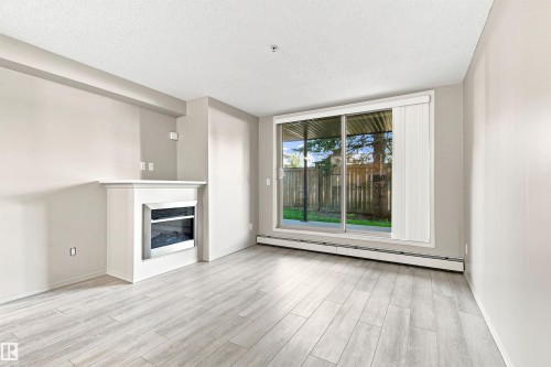 Light wood-finish flooring, built-in fireplace with a white mantel, sliding glass door, and vertical blinds - #117 17011 67 Av Nw, Edmonton, AB - Indoor Photo Showing Living Room With Fireplace