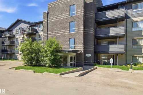 Multi-story building featuring a brick facade with horizontal banding, multiple balconies, an entrance with a covered overhang, and surrounding landscaping - #117 17011 67 Av Nw, Edmonton, AB - Outdoor With Facade