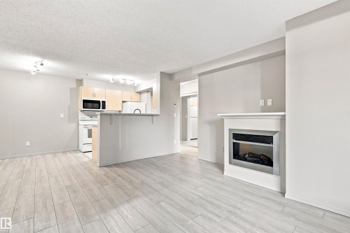 Modern fireplace with a white mantel, light wood-finish flooring, light-toned cabinetry, and white appliances - #117 17011 67 Av Nw, Edmonton, AB - Indoor With Fireplace