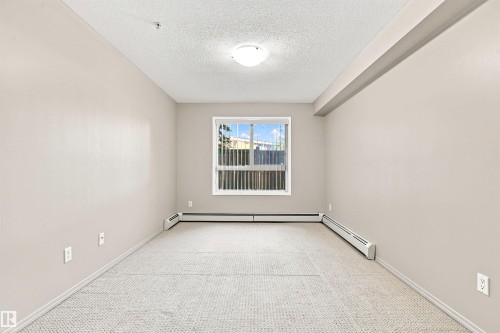 Neutral-toned wall paint, textured ceiling, window with vertical blinds, baseboard heating units, and light-toned carpet flooring - #117 17011 67 Av Nw, Edmonton, AB - Indoor Photo Showing Other Room