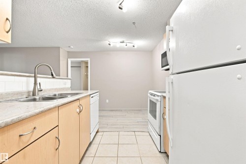 Kitchen featuring wood-finish cabinetry, a double basin sink with a gooseneck faucet, a white tile backsplash, and white appliances including a refrigerator, range, microwave, and dishwasher - #117 17011 67 Av Nw, Edmonton, AB - Indoor Photo Showing Kitchen With Double Sink