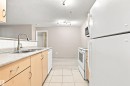 Kitchen featuring wood-finish cabinetry, a double basin sink with a gooseneck faucet, a white tile backsplash, and white appliances including a refrigerator, range, microwave, and dishwasher - #117 17011 67 Av Nw, Edmonton, AB  - Indoor Photo Showing Kitchen With Double Sink 
