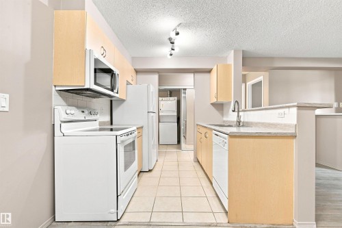 Galley kitchen featuring light wood-finish cabinetry, white appliances, and a tile backsplash - #117 17011 67 Av Nw, Edmonton, AB - Indoor Photo Showing Laundry Room