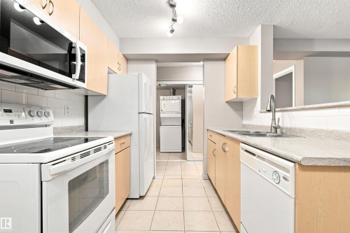 Galley kitchen with light wood-finish cabinetry, tile flooring, and white appliances including a refrigerator, range, microwave, and dishwasher - #117 17011 67 Av Nw, Edmonton, AB - Indoor Photo Showing Kitchen