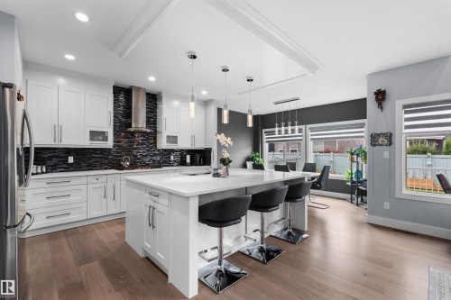Modern kitchen featuring white cabinetry, a large center island with seating, a black textured tile backsplash, stainless steel appliances, and wood-finish flooring - 16327 17 Avenue Sw, Edmonton, AB 