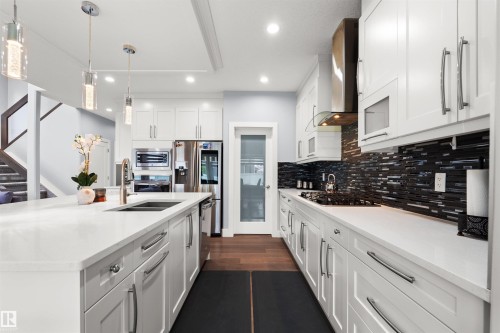Kitchen featuring white cabinetry, stainless steel appliances, dark wood-finish flooring, white countertops, and a dark mosaic tile backsplash - 16327 17 Avenue Sw, Edmonton, AB 
