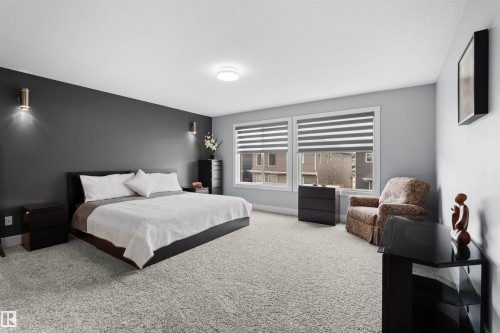 Carpeted bedroom featuring dual windows with horizontal blinds, a ceiling-mounted light fixture, and two wall-mounted sconces - 16327 17 Avenue Sw, Edmonton, AB 
