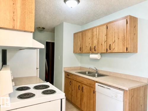 Kitchen featuring wood-finish cabinetry, a double basin stainless steel sink, a dishwasher, and a four-burner electric range with an overhead hood - #106 10620 104 St Nw, Edmonton, AB - Indoor Photo Showing Kitchen With Double Sink