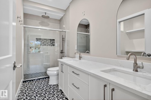 Spacious bathroom featuring a dual vanity with undermount sinks, patterned floor tiling, a glass-enclosed shower with a rainfall showerhead, and arch-top mirrors - 9736 73 Avenue Nw, Edmonton, AB 