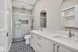 Spacious bathroom featuring a dual vanity with undermount sinks, patterned floor tiling, a glass-enclosed shower with a rainfall showerhead, and arch-top mirrors - 
