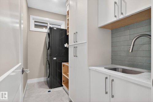 Laundry area featuring stacked appliances, white cabinetry with silver hardware, a utility sink with a gooseneck faucet, and a tiled backsplash - 9736 73 Avenue Nw, Edmonton, AB 