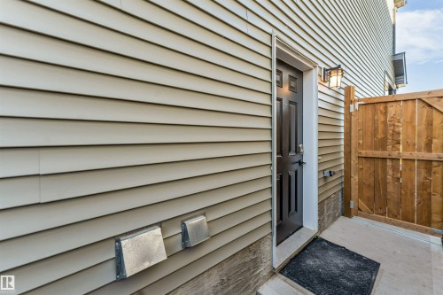 Beige horizontal siding, exterior door with paneling, wall-mounted exterior light fixture, concrete pathway, and wood fencing - 3236 Chernowski Way Sw, Edmonton, AB - Outdoor