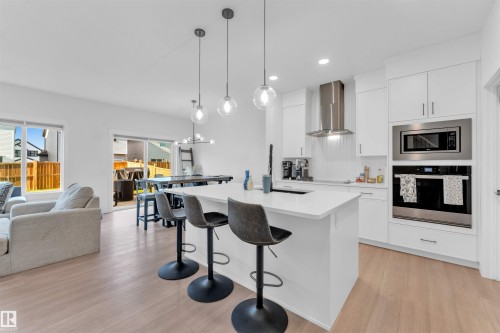 Open-concept living area with wood-finish flooring, white cabinetry, integrated stainless steel appliances, and a kitchen island with pendant lighting - 3236 Chernowski Way Sw, Edmonton, AB - Indoor Photo Showing Kitchen With Upgraded Kitchen